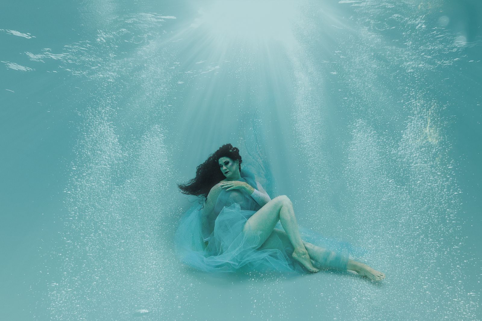 Fine-art underwater maternity portrait in soft blue tulle, captured by White Lavender Photography in Flower Mound, Texas — ethereal light beams and graceful pose create a dreamlike underwater atmosphere
