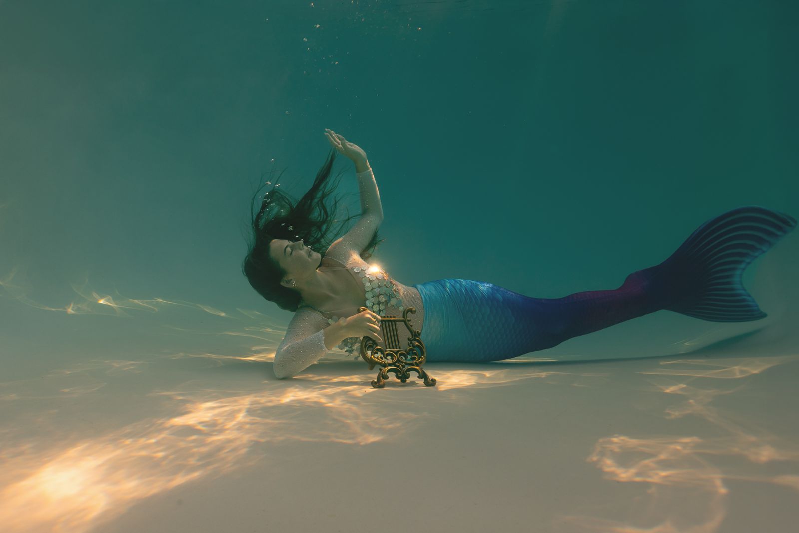 Artistic underwater mermaid portrait featuring a blue-tailed mermaid playing a golden harp beneath soft light reflections — fine-art photography by White Lavender Photography in Dallas, Texas
